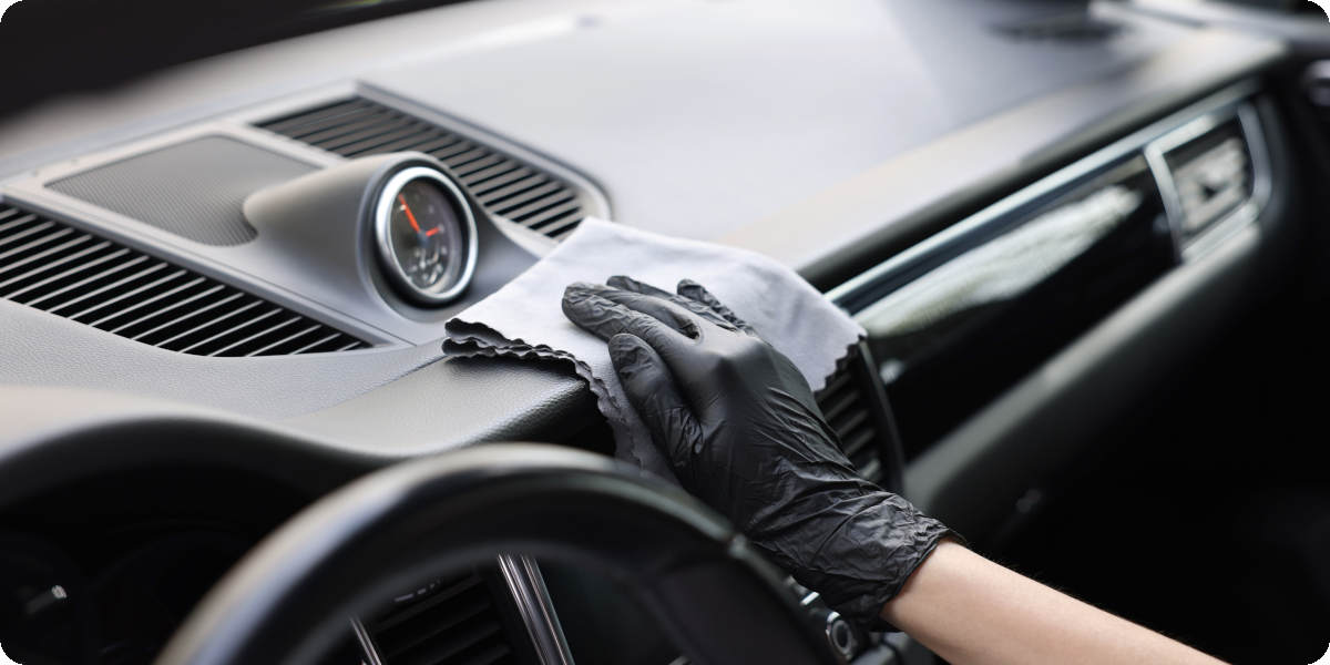 Eine Person tr&auml;gt schwarze Handschuhe und reinigt das Armaturenbrett eines Autos mit einem grauen Mikrofasertuch.