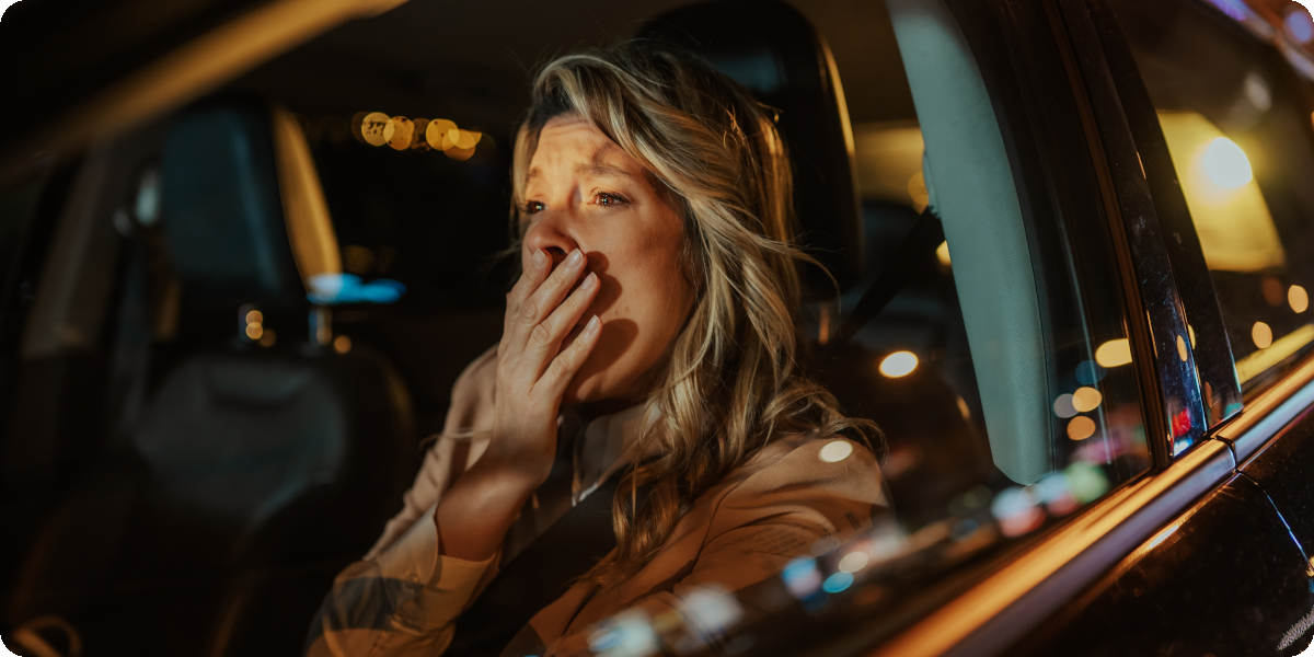 Eine blonde Frau sitzt in der Nacht am Steuer eines fahrenden Autos und hält sich die Hand vor dem Mund, weil sie gähnt.