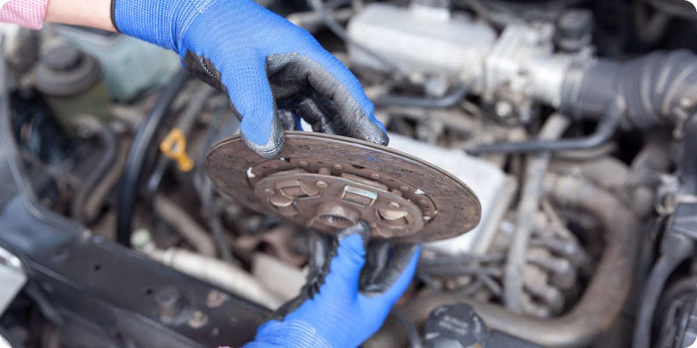 Zwei H&auml;nde mit blauen Gummihandschuhen halten eine verschlissene Kupplungsscheibe &uuml;ber dem Motorblock. 