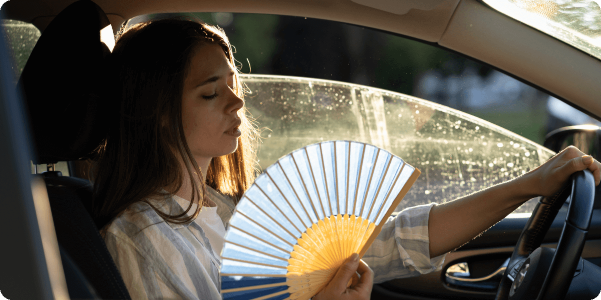Eine schwitzende Frau sitzt im Auto am Steuer und Fächert sich Luft ins Gesicht.