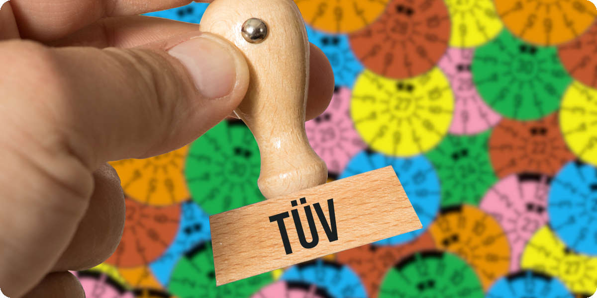 Eine Hand hält einen Tüv Stempel und im Hintergrund sind unterschiedliche Tüv Siegel zu sehen.