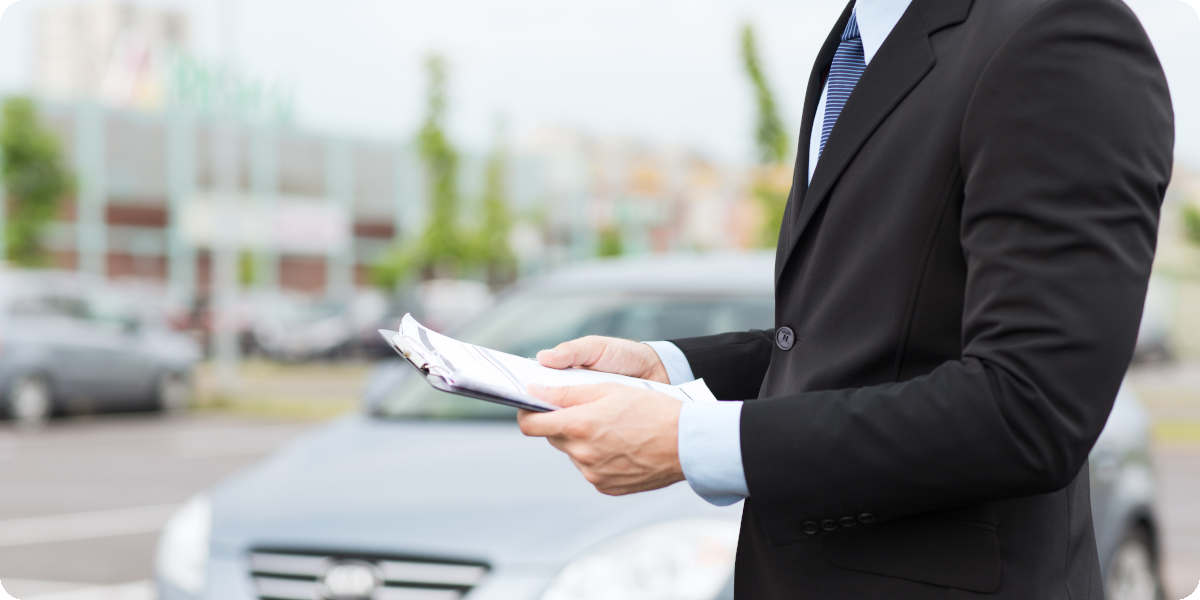 Autoverkäufer steht vor einem Auto und hält ein Klemmbrett mit einem Kaufvertrag in der Hand.