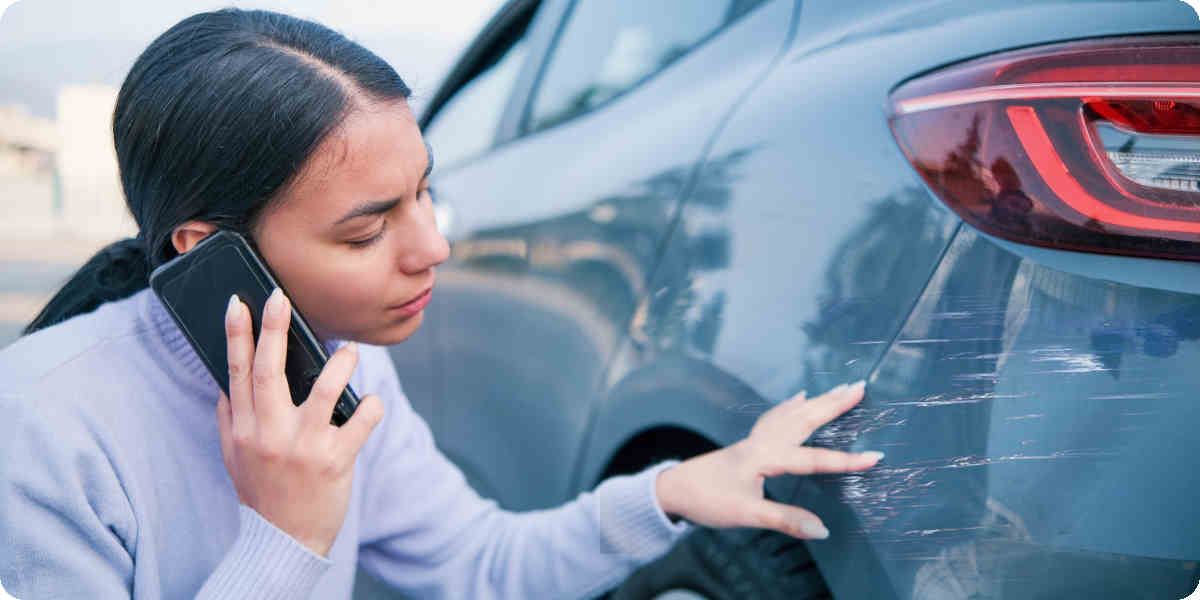 Eine Frau telefoniert mit der Polizei und begutachtet einen Parkrempler am Auto.