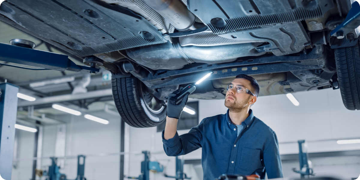 Prüfer vom TÜV sieht sich mit Taschenlampe den Unterboden eines Autos an. 