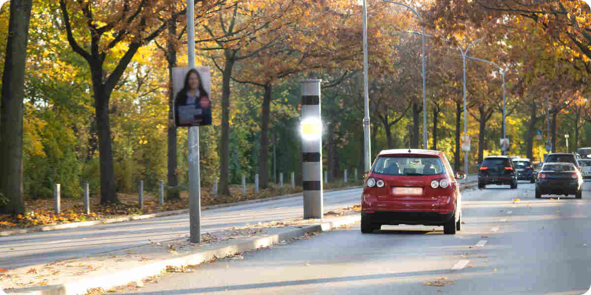 Ein rotes Auto wird auf einer 3 spurigen Straße in Deutschland von einem Standblitzer geblitzt.