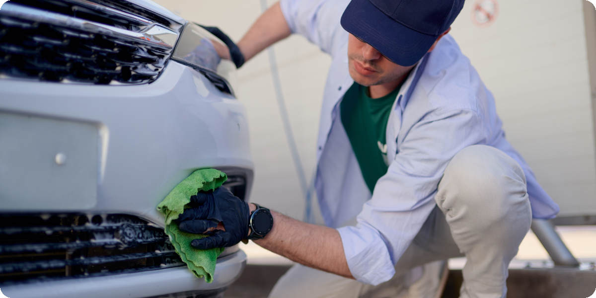 Eine Person reinigt die vordere Stoßstange eines Autos mit einem grünen Mikrofasertuch.
