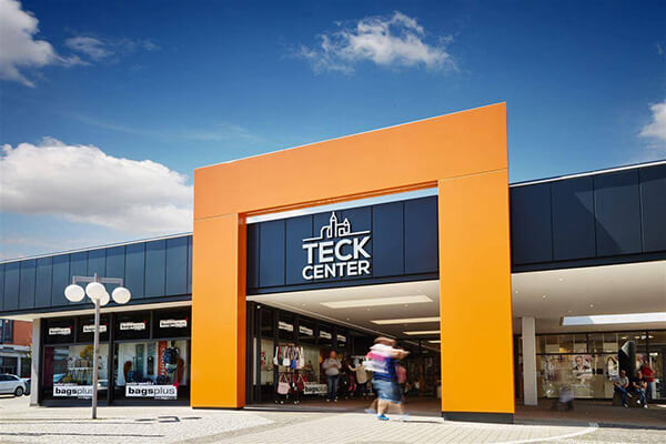 Eingang des Teck Centers mit markantem orangefarbenem Portal und Geschäften wie BagsPlus unter blauem Himmel in Kirchheim unter Teck