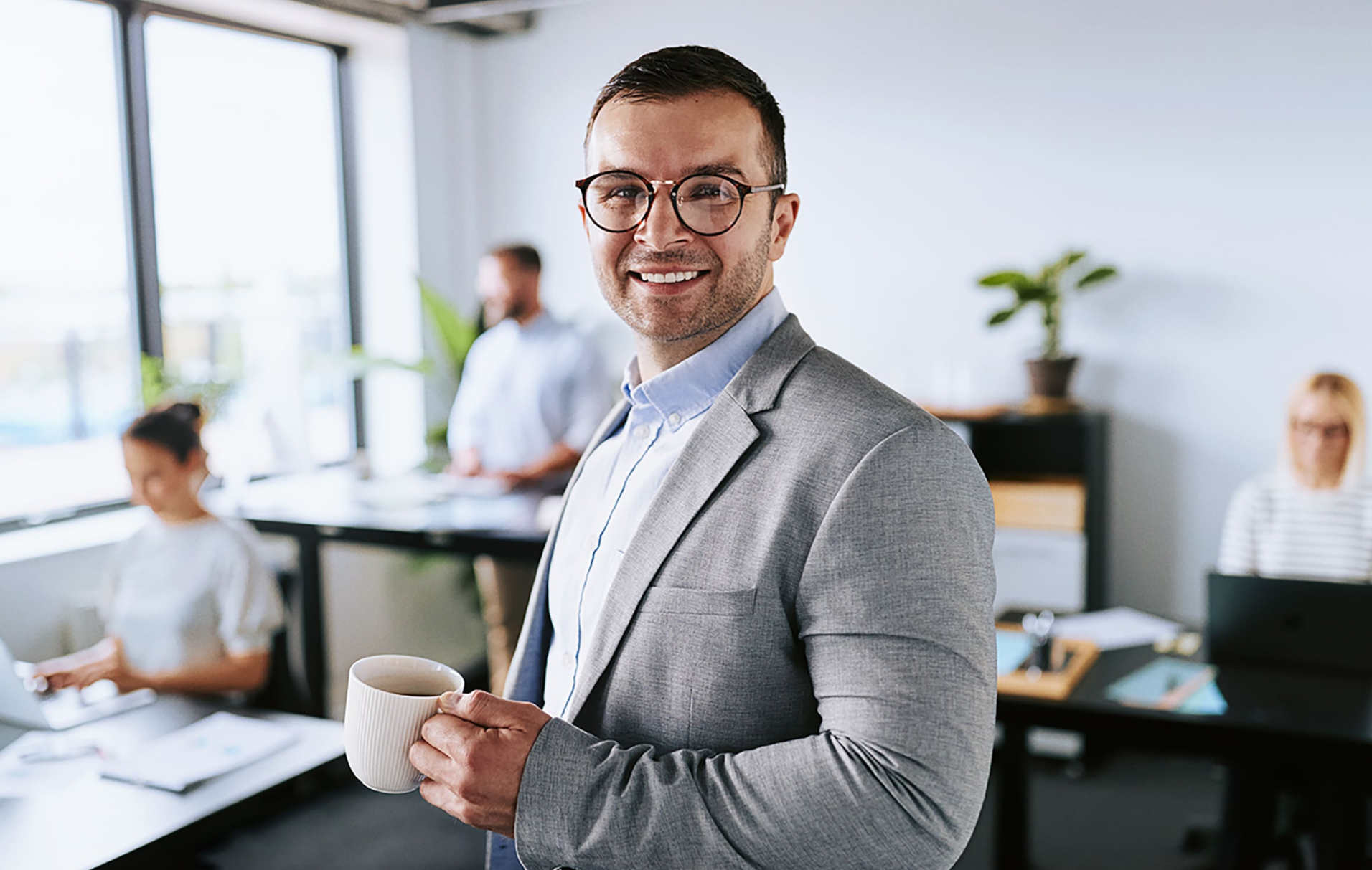Ein Mann im Anzug hält im Büro eine Tasse in der Hand und lächelt in die Kamera.