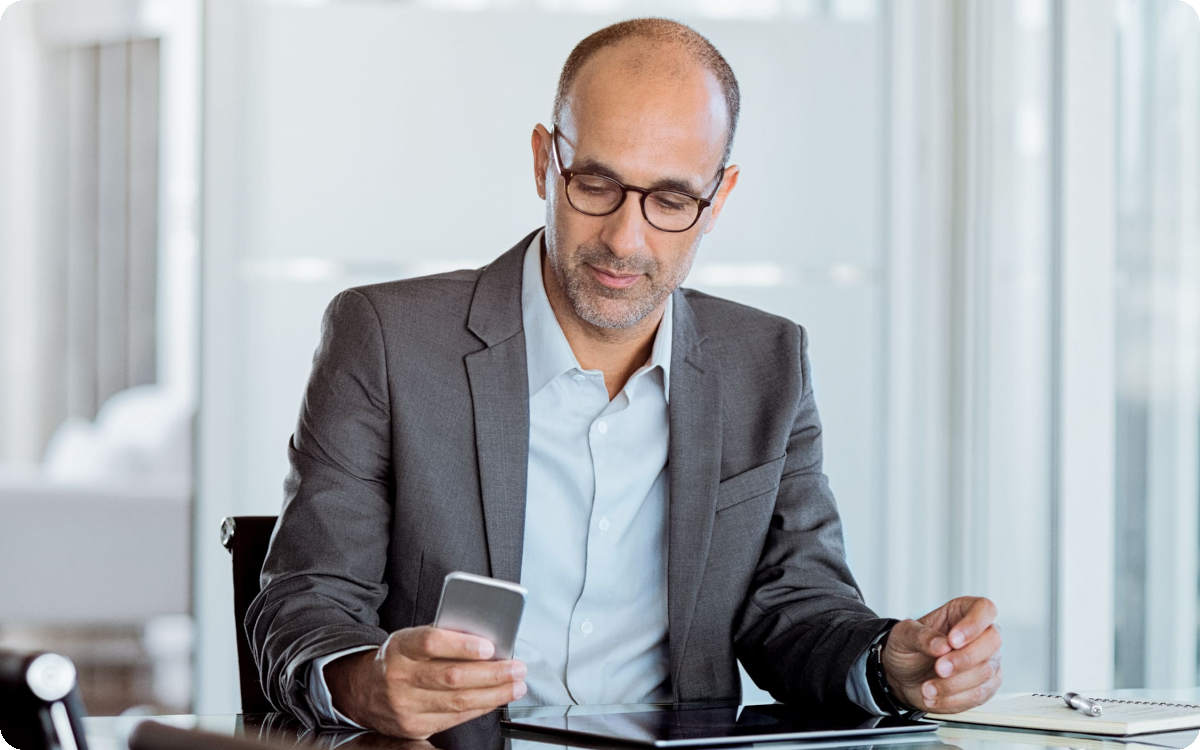 Ein Mann im Anzug sitzt am Schreibtisch und schaut auf sein Smartphone.