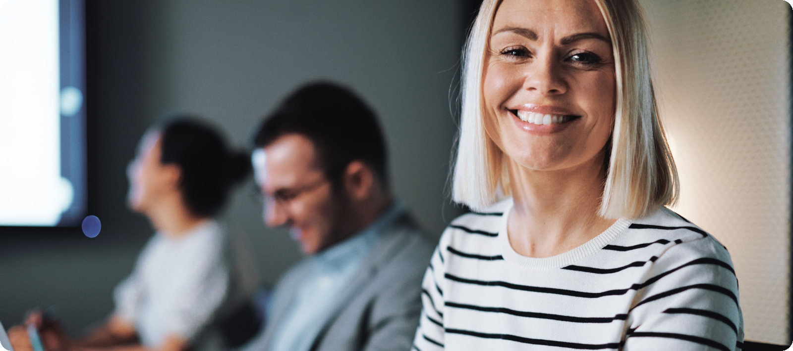 Eine blonde Frau sitzt im Büro und lächelt in die Kamera.